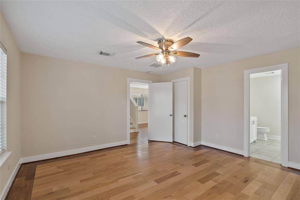 This photo shows a bright, spacious bedroom with hardwood floors and neutral walls. It features a ceiling fan, two doors, and an adjoining bathroom. Large windows provide ample natural light.