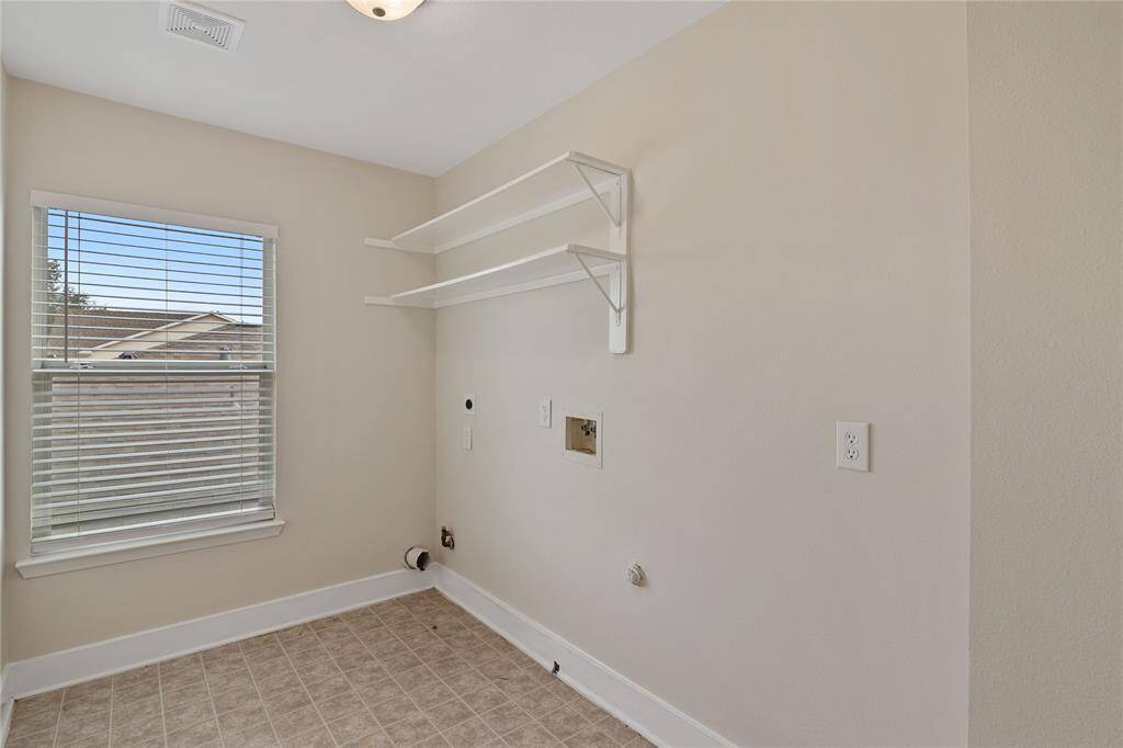 This photo shows a laundry room with hookups for a washer and dryer, featuring a window for natural light, tiled flooring, and two wall-mounted shelves for storage.
