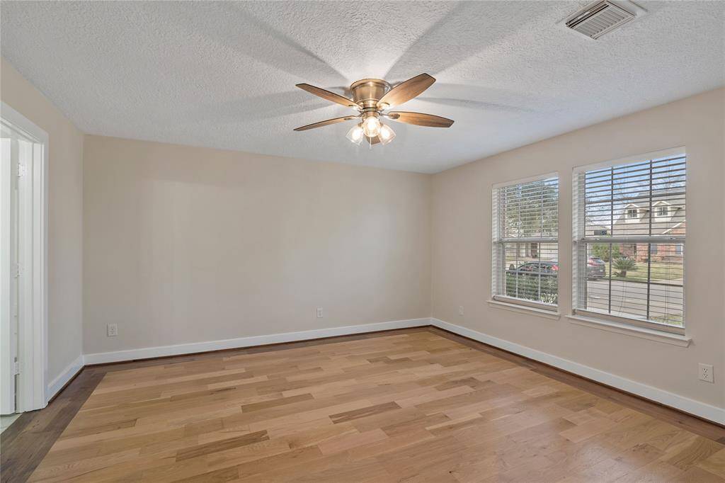 This bright room features hardwood floors, a ceiling fan with lighting, and large windows that provide ample natural light. The neutral walls offer a versatile space ready for personalization.