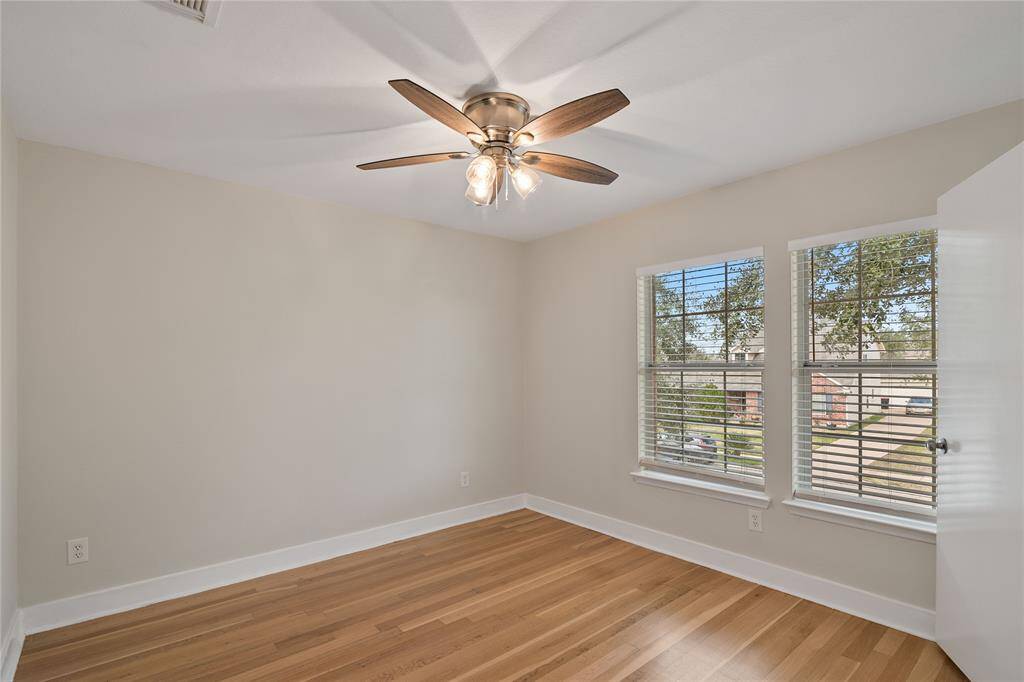 This room features hardwood flooring, neutral walls, and a ceiling fan with lights. It has two large windows that let in ample natural light, creating a bright and airy feel.