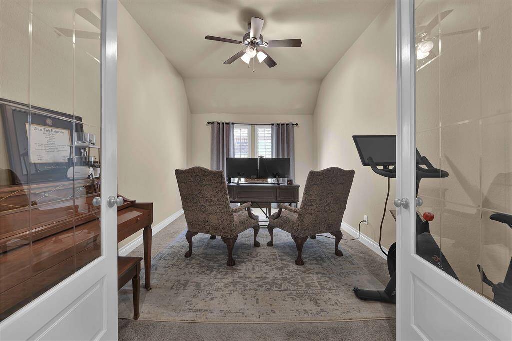 The private home office offers a dedicated space for work or study, featuring a high ceiling with a ceiling fan and light kit for added comfort. Large windows adorned with plantation shutters fill the room with natural light, while plush carpet underfoot creates a cozy atmosphere.