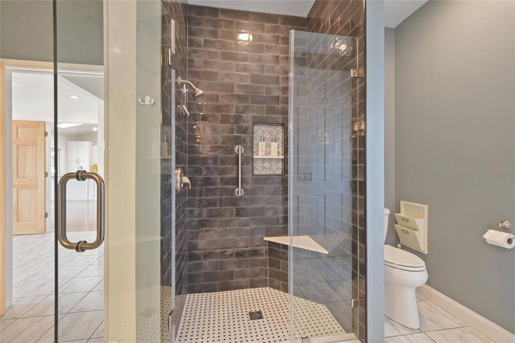 Across from the vanity area is this generously sized, renovated shower with seamless glass door, updated designer tile, shelf for shampoo & conditioner, grab bar for safety, and seat.  To the right is the discrete alcove for the commode.