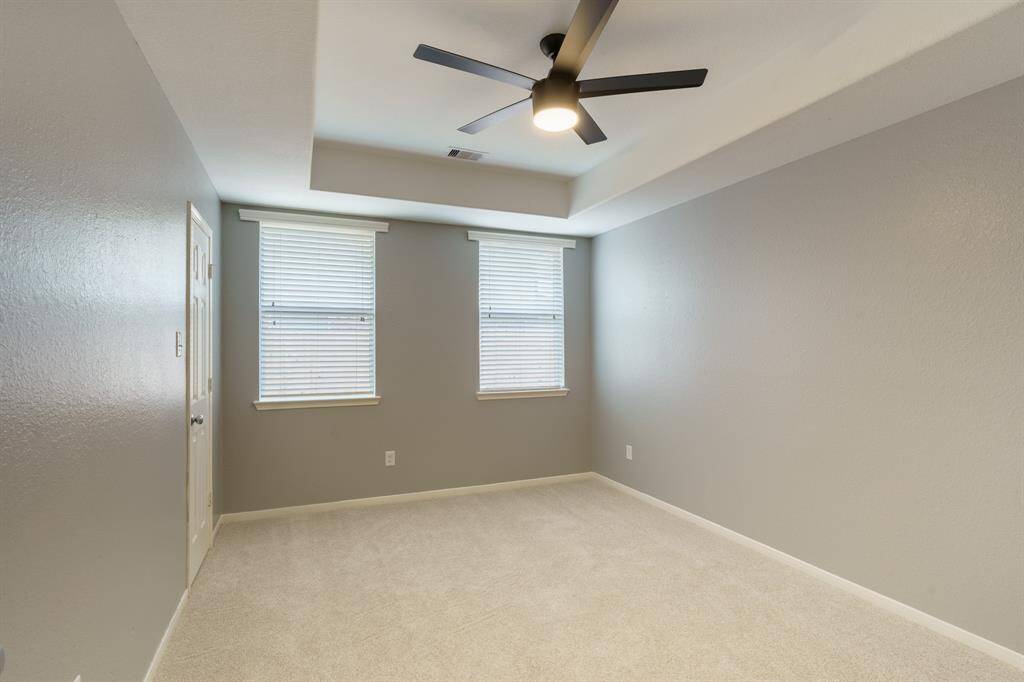 This room features neutral walls, carpeted flooring, and a modern ceiling fan. It has two windows with blinds, offering natural light, and a simple yet elegant tray ceiling design.