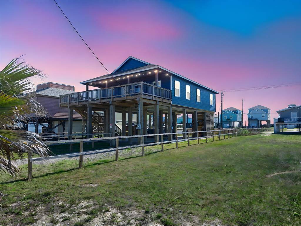 Elevated navy blue beach house with a spacious partially covered wooden deck, situated on stilts for coastal living. It features a modern blue exterior, ample parking or storage space underneath, and is surrounded by a fenced grassy area. Ideal for enjoying gulf and bay views and breezy outdoor relaxation.