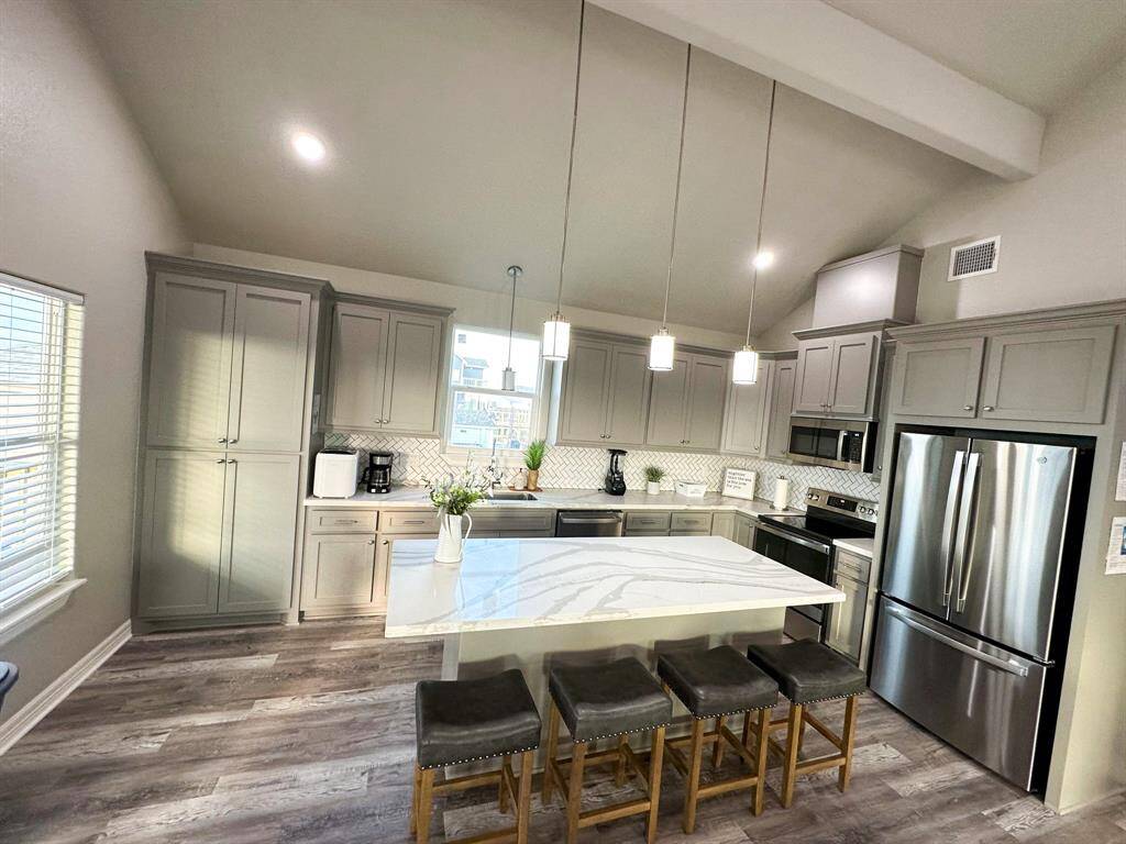 This kitchen features modern gray cabinets, a spacious marble island with seating for four, stainless steel appliances, and elegant pendant lighting, all under a vaulted ceiling for an airy feel.