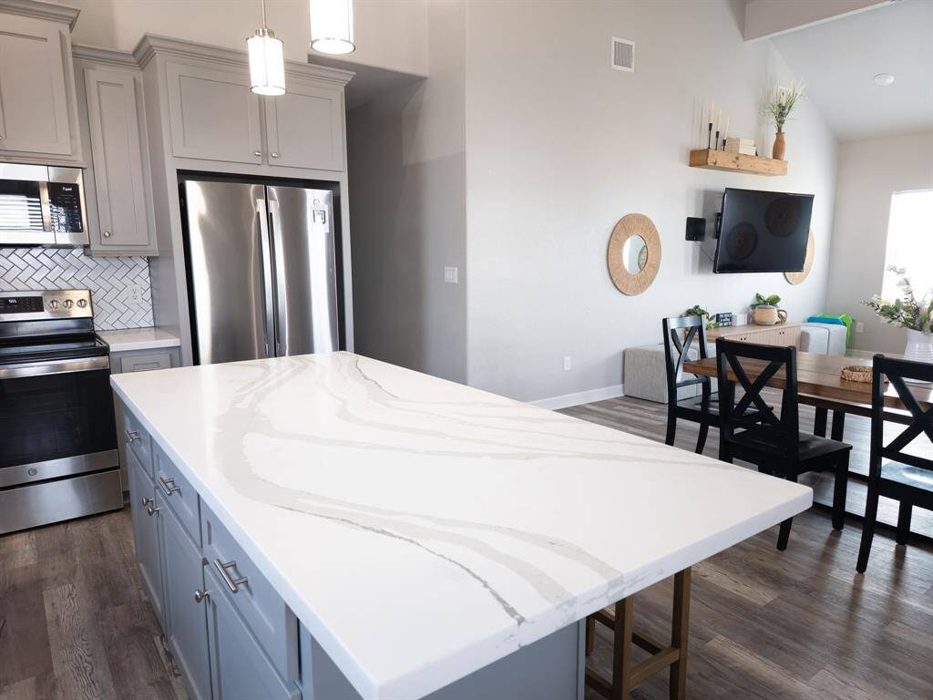This modern kitchen features a sleek quartz island with a marble-style countertop, stainless steel appliances, and cabinetry. Adjacent is an open dining/living area with a wall-mounted TV and contemporary decor, creating a bright and inviting space.