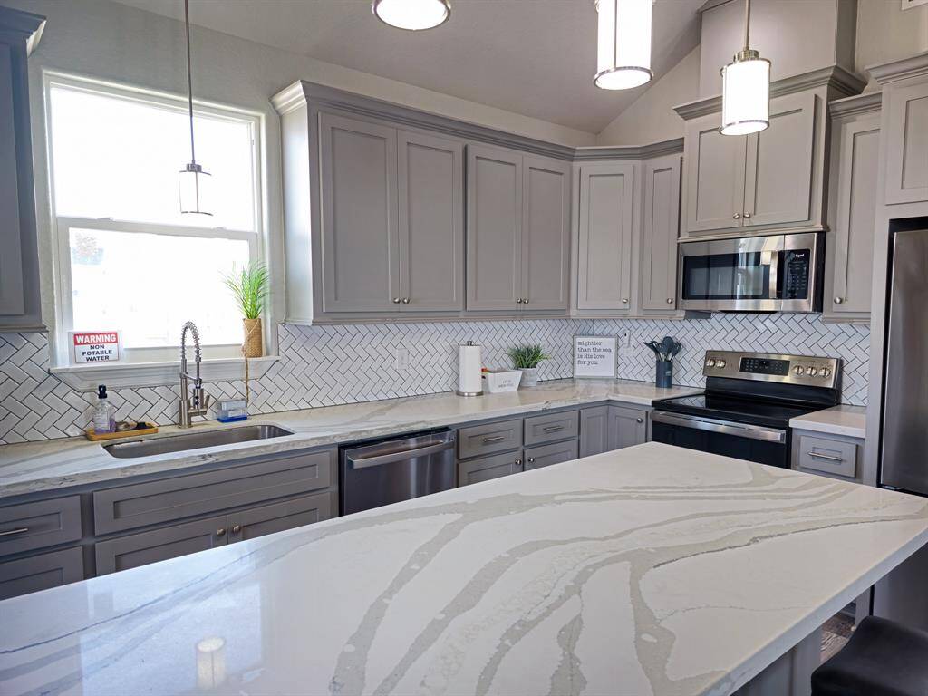 This kitchen features modern cabinetry, stainless steel finish appliances, a spacious marble-patterned quartz island, and a stylish herringbone backsplash. Natural light enhances the open, airy feel, making it ideal for cooking and entertaining.
