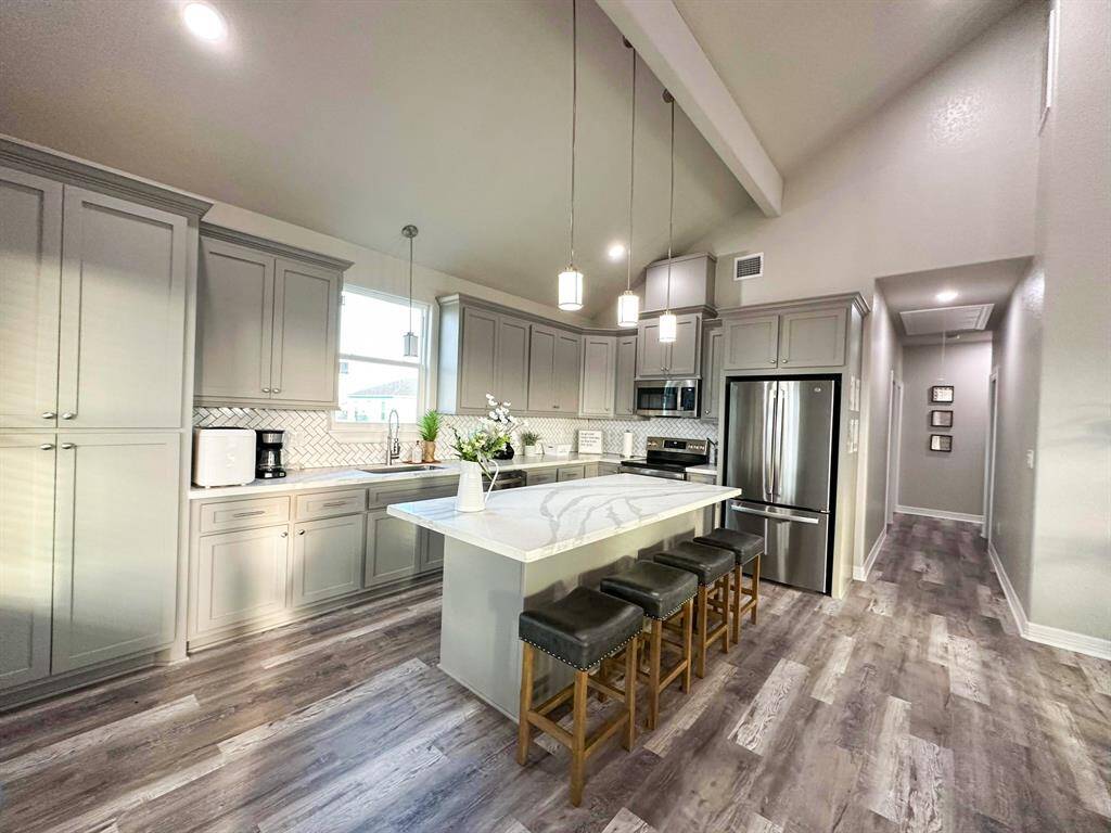 The kitchen with a spacious island with seating, stainless steel finish appliances, and elegant pendant lighting. The space is complemented by a light floor and a stylish backsplash, creating a bright and inviting atmosphere.