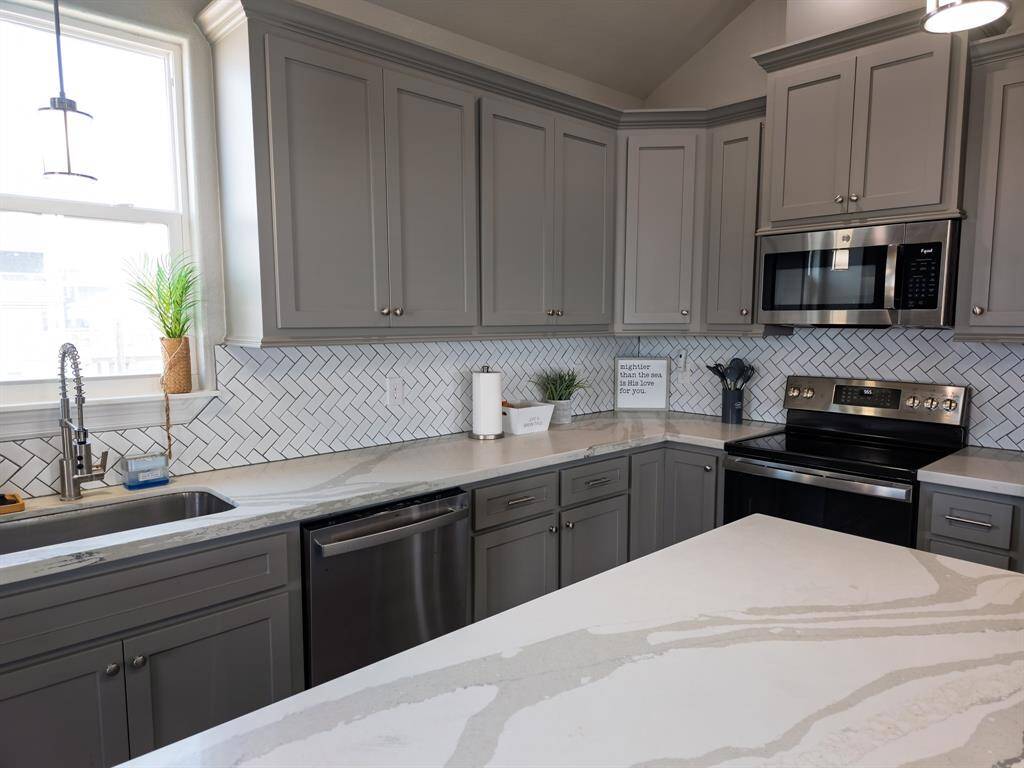 This kitchen features elegant shaker cabinetry with modern stainless steel finish appliances. The countertops are a sleek, white marble pattern, complemented by a stylish herringbone tile backsplash. Bright and inviting, it includes a large window and an island for extra entertaining space.