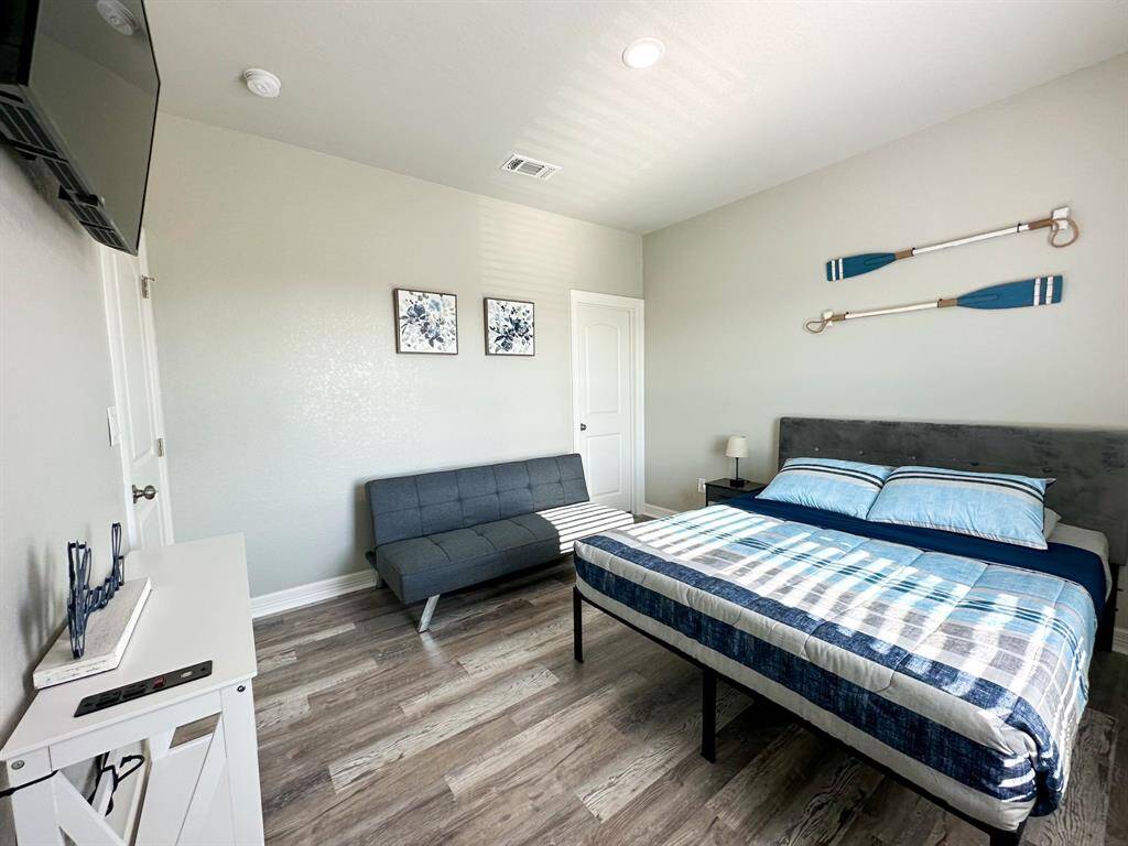 This bedroom features a cozy double bed with nautical-themed decor, including oars on the wall. It has a gray futon, wood-look flooring, and a wall-mounted TV for comfort and entertainment and comes with en-suite. The room is bright and inviting with simple, modern furnishings.