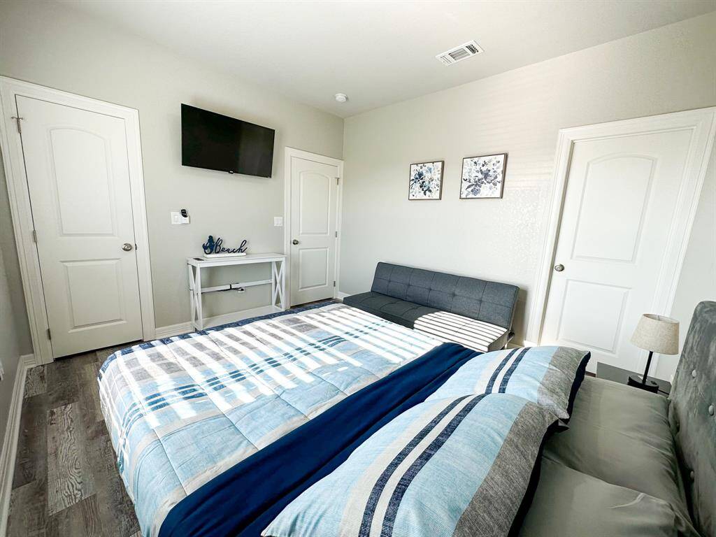 This cozy bedroom features a stylish bed with blue and gray bedding, a wall-mounted TV, and a small gray sofa. The room is accented with floral artwork and has ample natural light from a window. Dual white doors add to the clean, modern aesthetic.