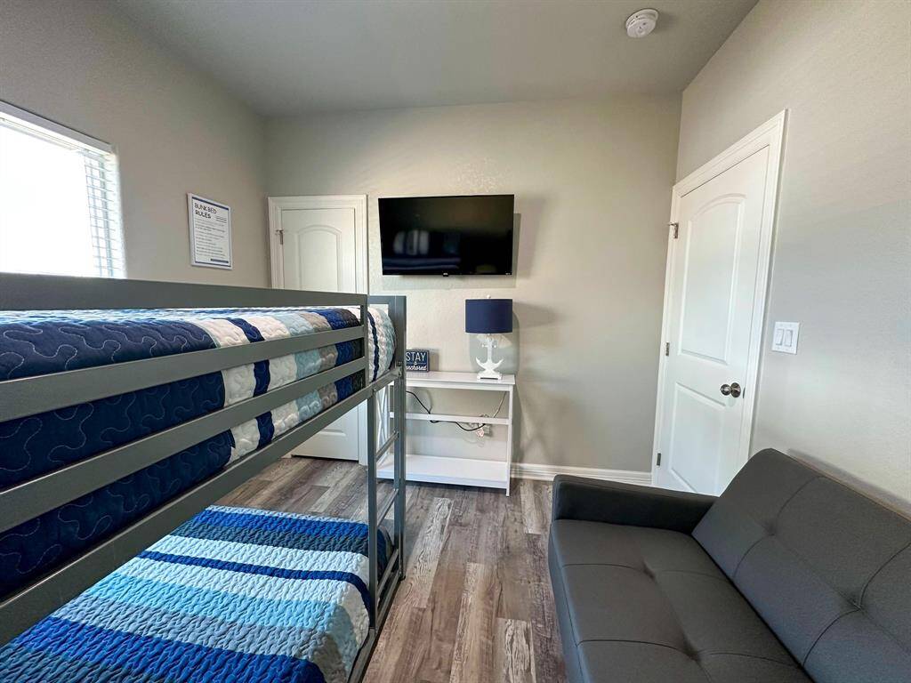 This bedroom features a bunk bed with colorful bedding, a wall-mounted TV, and a small sofa. The room has laminate wood flooring and neutral walls, creating a cozy and functional space.