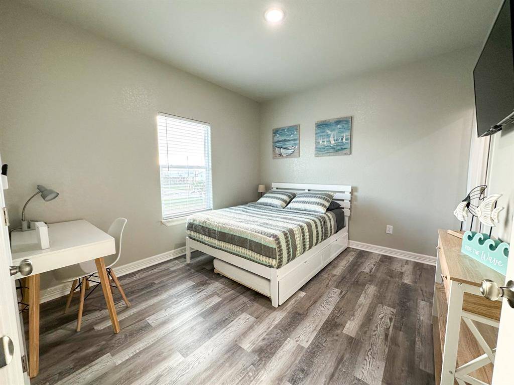 This bedroom features a neatly made bed with striped bedding, a desk with a lamp, a window that lets in natural light and an ensuite! The walls are painted a soft neutral color, and the room has stylish wood style flooring. It’s decorated with nautical-themed artwork and a small TV mounted on the wall.