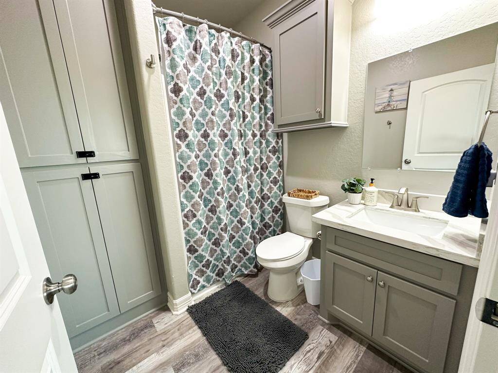 The 1st of 3 ensuite baths features modern gray cabinetry, a shower with a patterned curtain, a stylish vanity with a sleek faucet, and wood-look flooring, offering a clean and contemporary feel.