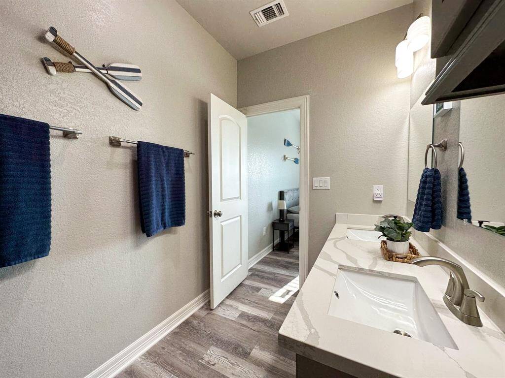 This 2nd ensuite bathroom features a modern design with a double sink vanity, sleek fixtures, and a large mirror. The neutral walls are complemented by dark blue towels and decorative accents. Light wood-look flooring adds warmth, and natural light streams in from an adjacent room.
