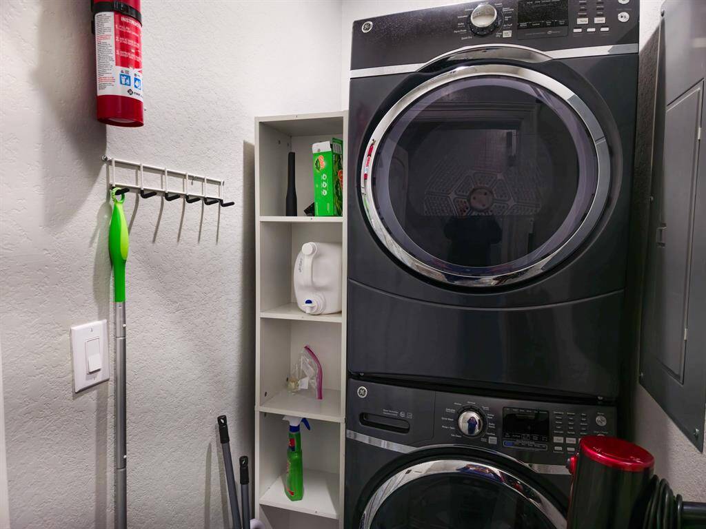 This compact laundry space features a modern stackable washer and dryer. It includes convenient shelving for storage, a wall-mounted fire extinguisher, and hooks for cleaning tools.