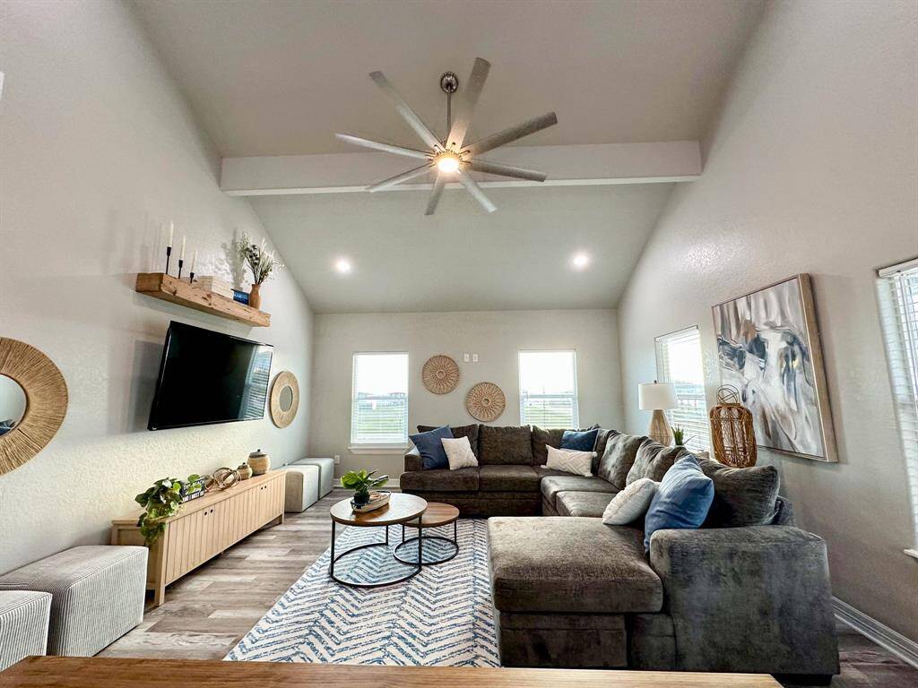 Spacious living room with high ceilings and abundant natural light. Features a large sectional sofa, modern ceiling fan, and mounted TV. Stylish decor includes wall art, a patterned rug, and a wooden shelf with plants. Ideal for comfortable living and entertaining.
