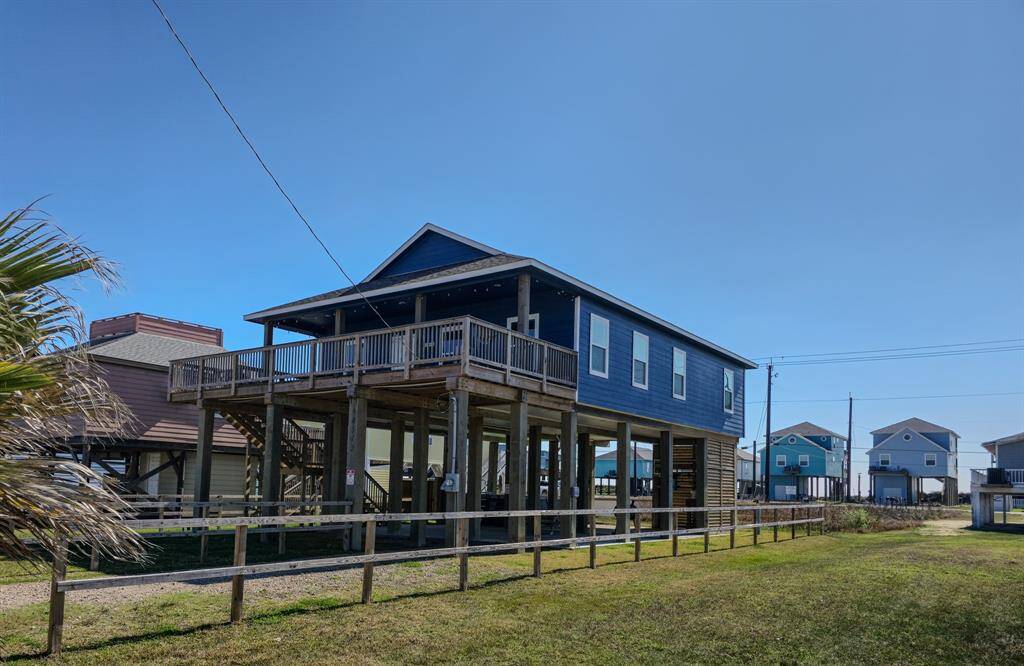 Elevated navy blue beach house with a spacious partially covered wooden deck, situated on stilts for coastal living. It features a modern blue exterior, ample parking or storage space underneath, and is surrounded by a fenced grassy area. Ideal for enjoying gulf and bay views and breezy outdoor relaxation.