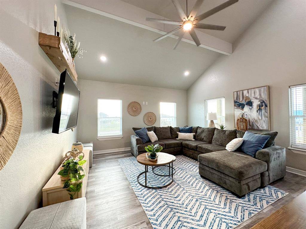 This spacious living room features high ceilings with a large fan, ample natural light from multiple windows, and a cozy sectional sofa. It includes a wall-mounted TV, stylish decor, and a patterned area rug, creating a modern and inviting space.