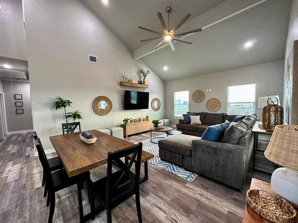 This photo showcases a modern, open-concept living and dining area with high ceilings. The space features stylish decor, a large sectional sofa, a wooden dining table, and a wall-mounted TV. Natural light fills the room through two windows, and the floor is covered with wood-style planks, creating a warm and inviting atmosphere.