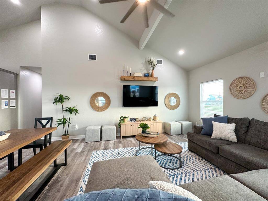 This spacious living room features high ceilings and abundant natural light. It includes a modern sectional sofa, a mounted TV, and stylish decor. The open layout seamlessly connects to a dining area with a wooden table and benches, creating a welcoming and functional space.