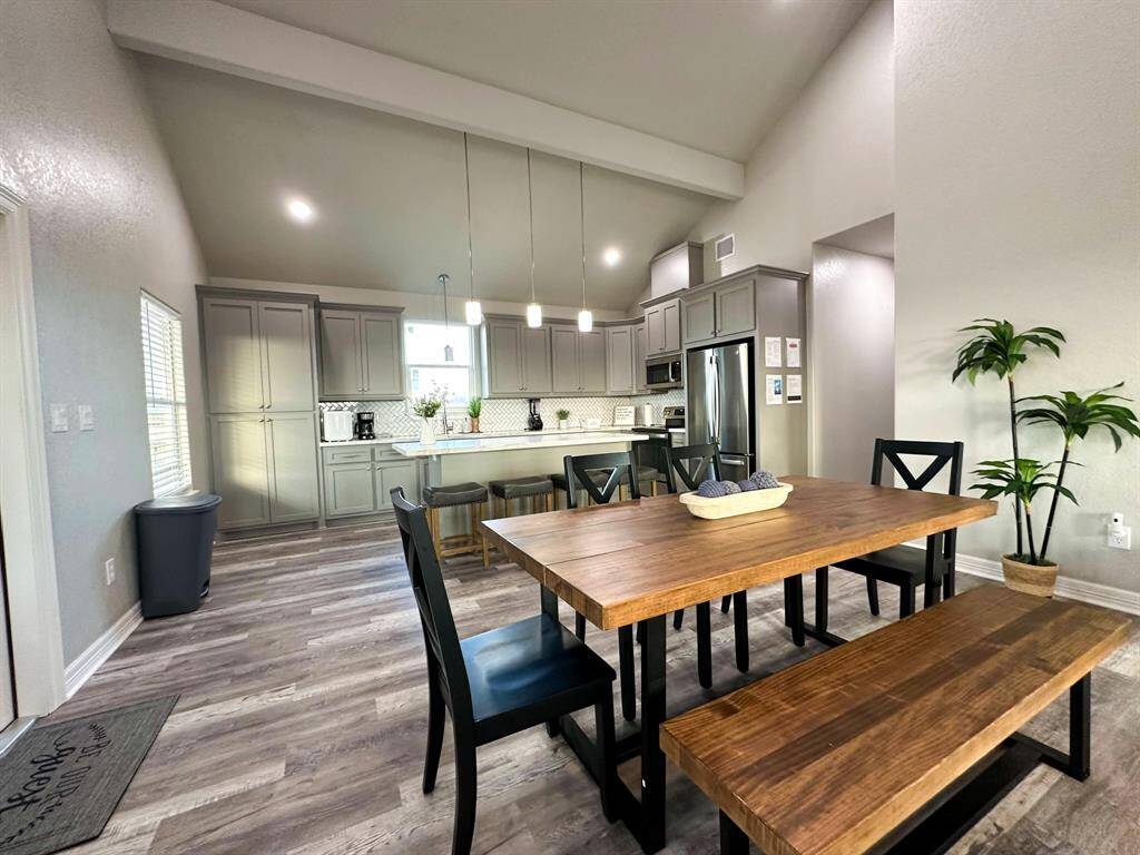 This photo showcases a modern, open-concept kitchen and dining area. The kitchen features sleek gray cabinetry, stainless steel appliances, and a central island with pendant lighting. The dining space includes a wooden table with bench seating, complemented by neutral walls and wood-style flooring for a warm, inviting atmosphere.