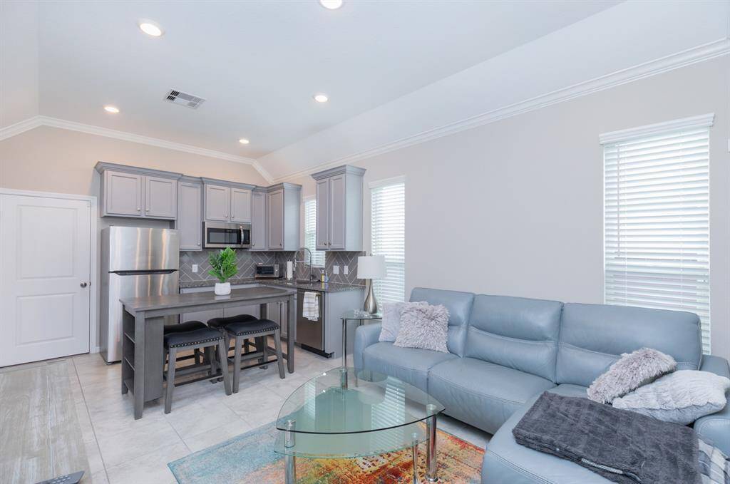 This photo highlights the stylish Next Gen suite, featuring a modern, open-concept living room and kitchen. The space includes sleek gray cabinetry, stainless steel appliances, and a cozy seating area with a light blue sofa adorned with decorative pillows. Large windows allow natural light to fill the room, creating a bright and welcoming atmosphere. Perfect for multi-generational living, this suite offers both comfort and convenience.