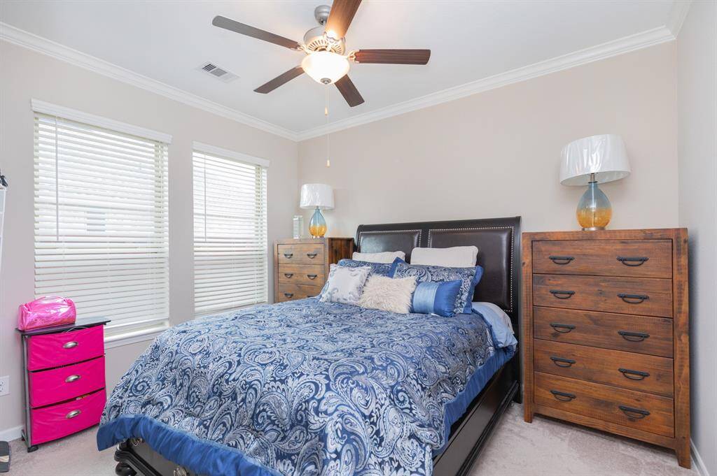 This cozy bedroom in the Next Gen suite offers a peaceful retreat with neutral walls, carpeted flooring, and a ceiling fan for added comfort. A large bed with a blue patterned comforter is complemented by two wooden dressers, providing ample storage space. The room is bathed in natural light from two windows, creating a serene atmosphere perfect for rest and relaxation.