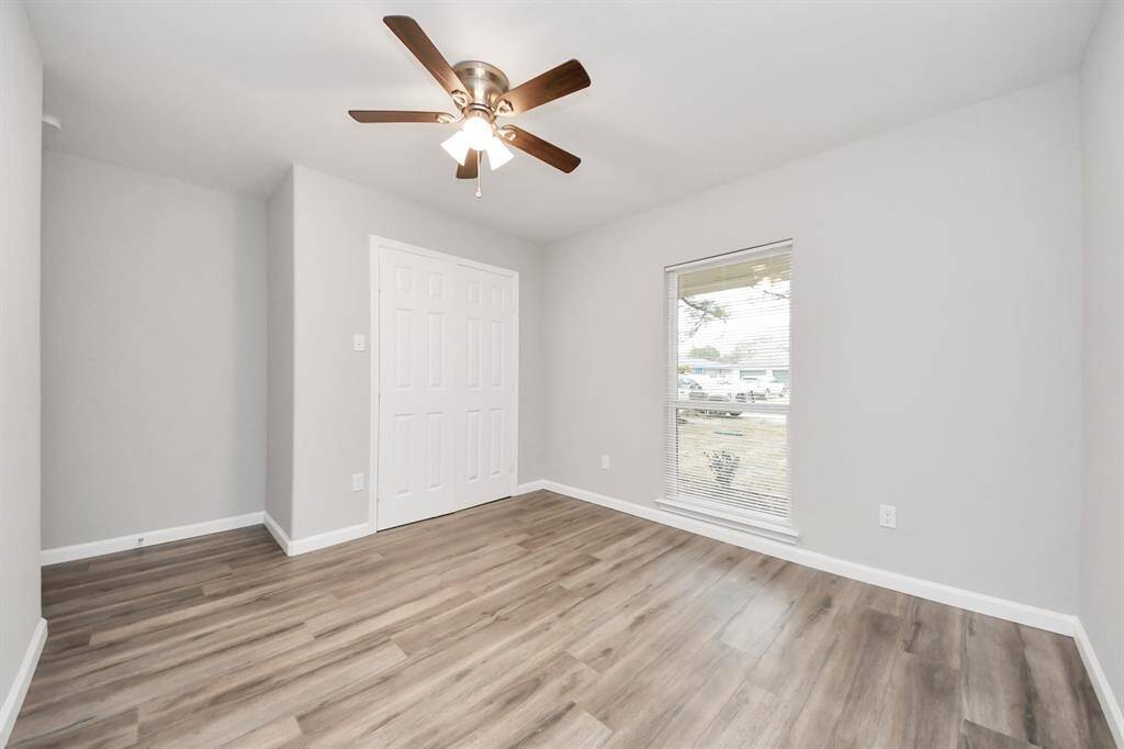 This room features a modern design with light gray walls laminate wood flooring. It has a ceiling fan for comfort and a large window for natural light. A double-door closet provides ample storage space.