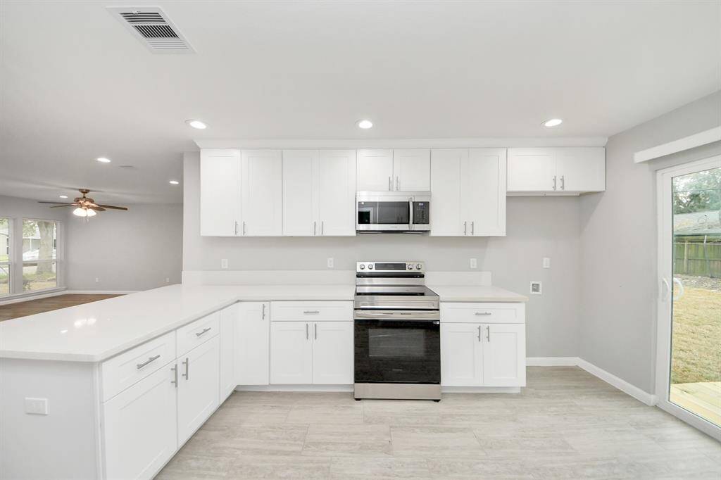 This modern kitchen features sleek white cabinetry, stainless steel appliances, and a spacious countertop. It has an open layout with a view to the living area and access to the backyard through sliding glass doors, offering plenty of natural light.