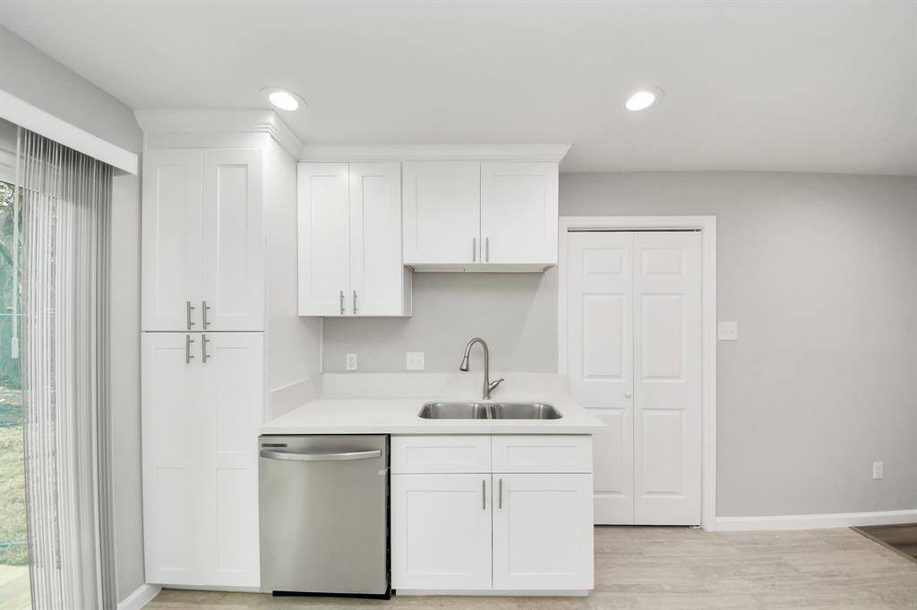This photo showcases a modern kitchen area with sleek white cabinetry, a stainless steel dishwasher, and a double sink. The room is well-lit with recessed lighting and has a clean, contemporary design. Sliding glass doors offer natural light and access to the outdoors.