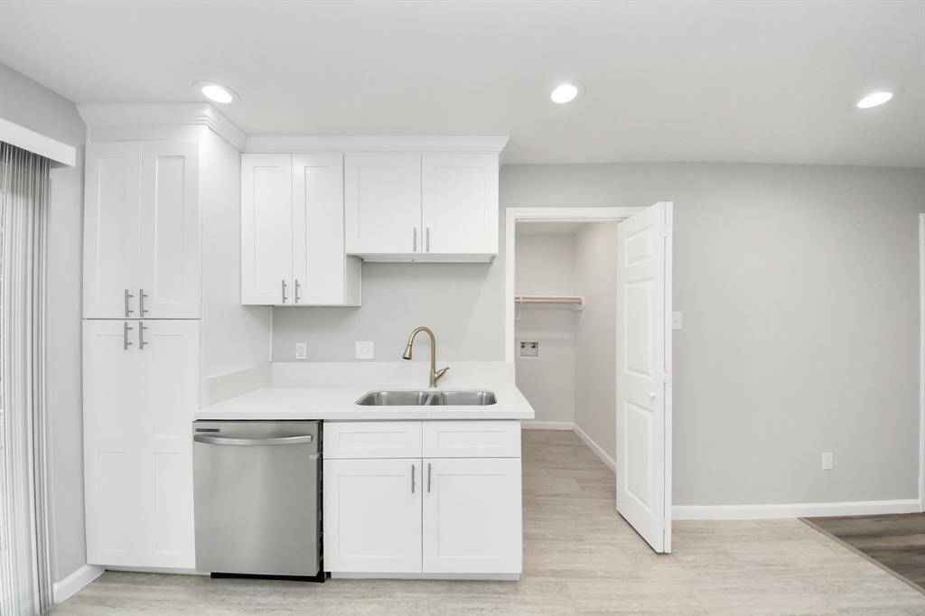 This photo showcases a modern kitchen with sleek white cabinetry, stainless steel dishwasher, and a double sink. The space is bright with recessed lighting, and there's an adjacent utility room with a door. The neutral tones create a clean and inviting atmosphere.