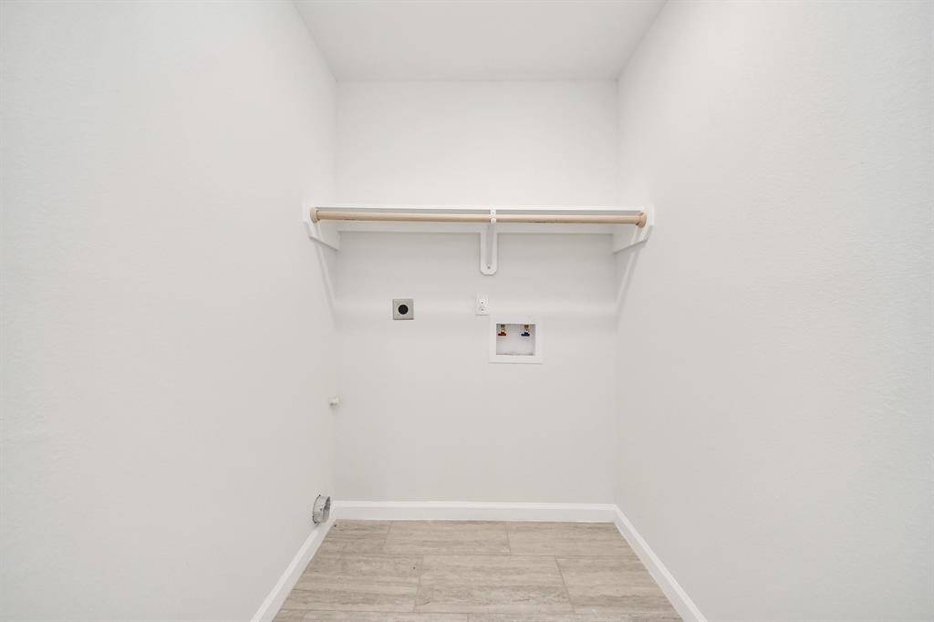 This is a small laundry room with white walls, light wood flooring, and plumbing hookups for a washer and dryer, featuring a wooden shelf for storage.