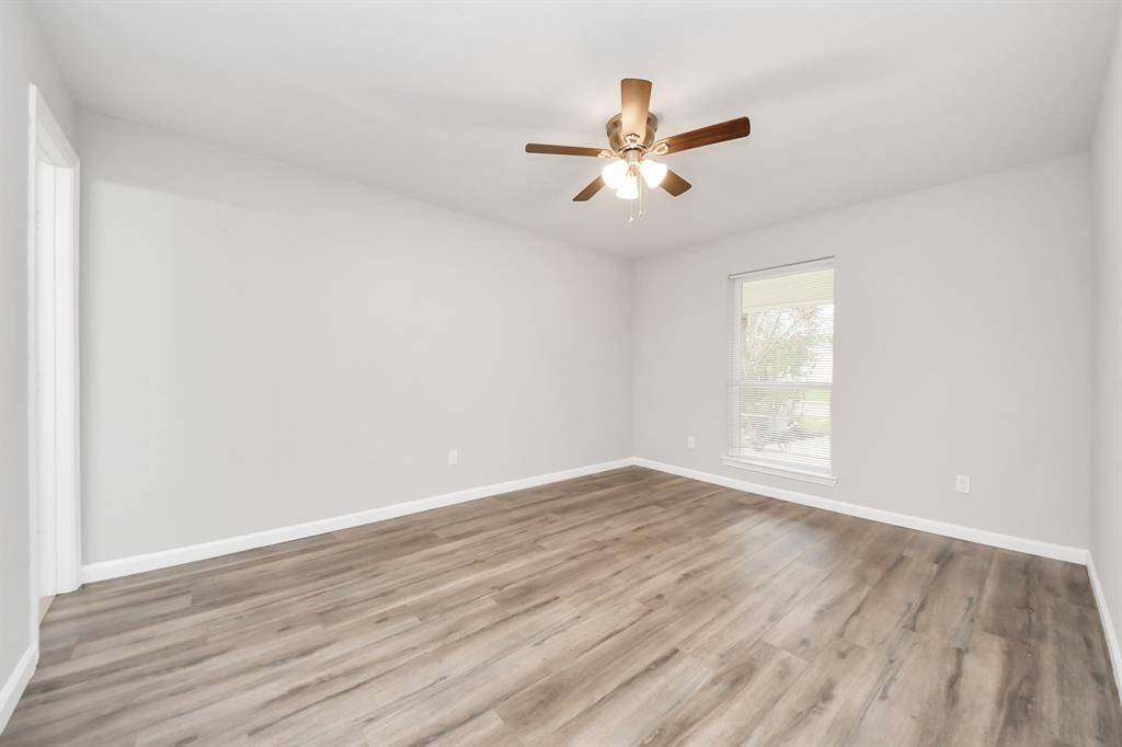 This room features a clean, modern design with light gray walls and wood-look flooring. It includes a ceiling fan for comfort and a window allowing natural light to brighten the space.