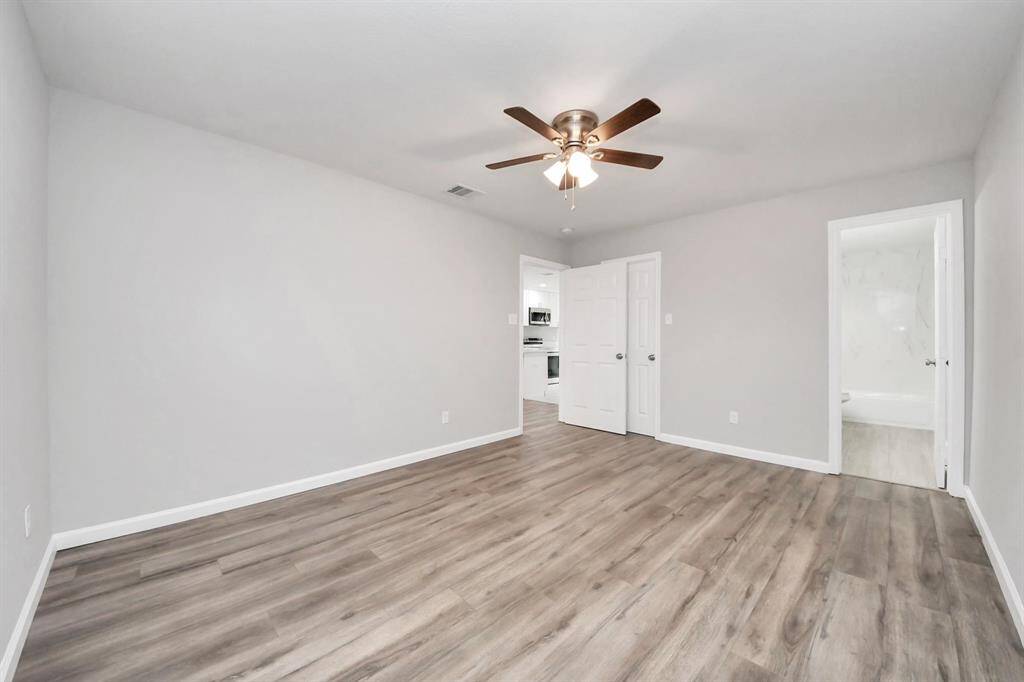 This spacious room features light gray walls, laminate wood-style flooring, and a ceiling fan. It connects to a kitchen area through white double doors and has an adjacent bathroom with a tub.