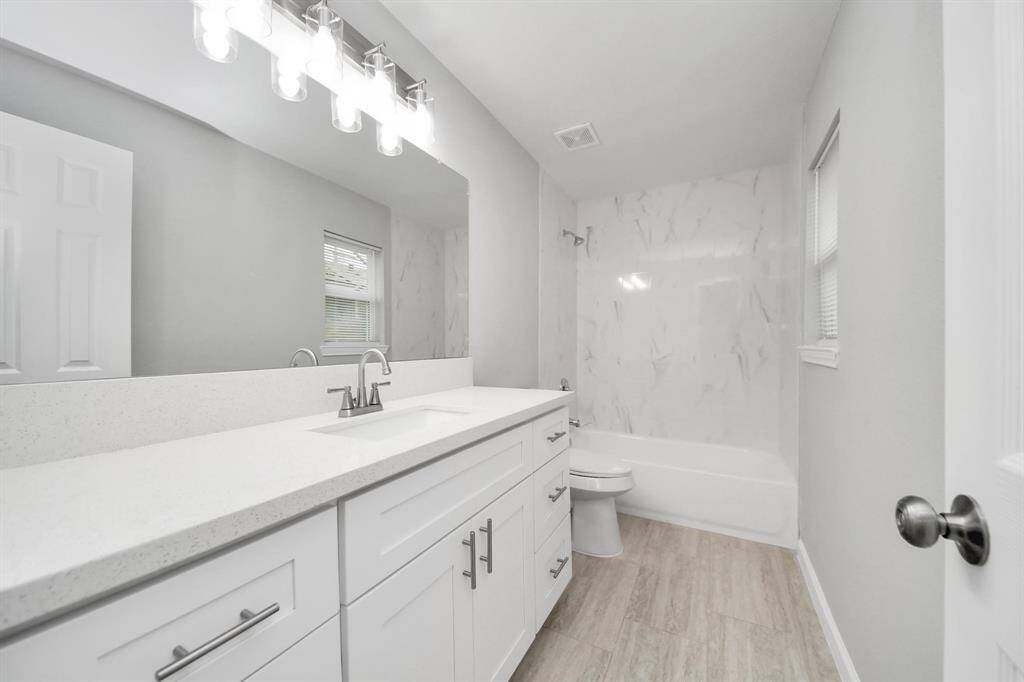 This bathroom features a modern design with light gray walls, a spacious white vanity with ample storage, a large mirror, and sleek lighting fixtures. The bathtub area is accented with elegant patterned tiles, and the floor has a stylish wood-look finish.