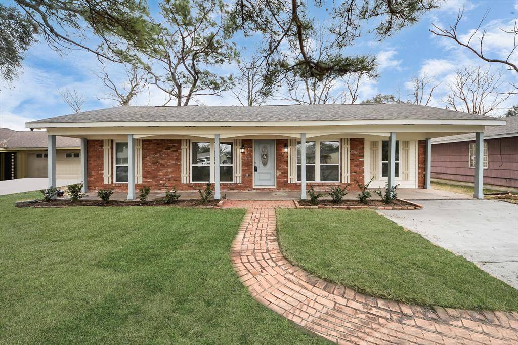 This charming single-story brick home features a welcoming front porch with wide windows, a neatly manicured lawn, and a brick walkway leading to the entrance. The attached carport adds convenience and enhances curb appeal.