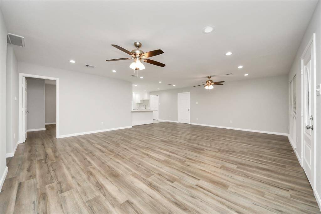 This spacious open-concept living area features modern laminate wood flooring, neutral walls, recessed lighting, and ceiling fans. It seamlessly connects to a white kitchen, creating an inviting, flexible space ideal for entertaining.
