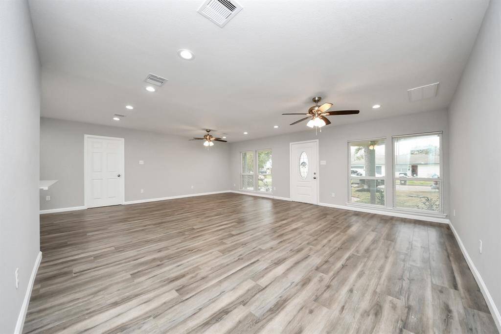 This spacious living room features modern laminate wood flooring, neutral walls, recessed lighting, and ceiling fans. Large windows provide plenty of natural light, and the front door has a decorative glass panel.