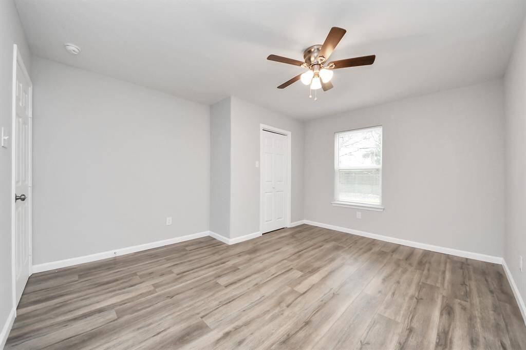 This room features fresh gray walls, laminate wood flooring, and a ceiling fan with lights. It includes a window for natural light and a white closet door, offering a clean and modern look.