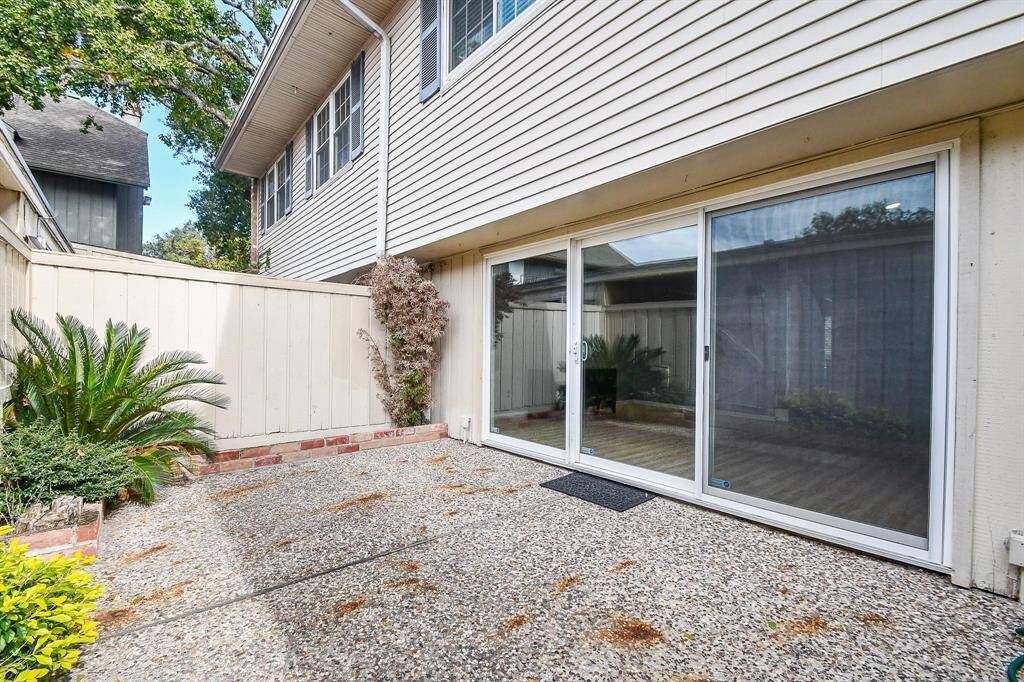 This photo showcases a private outdoor patio area with a pebbled surface, bordered by a low brick and white wooden fence. It's adjacent to a house with sliding glass doors, offering easy indoor-outdoor access. Lush greenery adds a touch of nature to the cozy space.