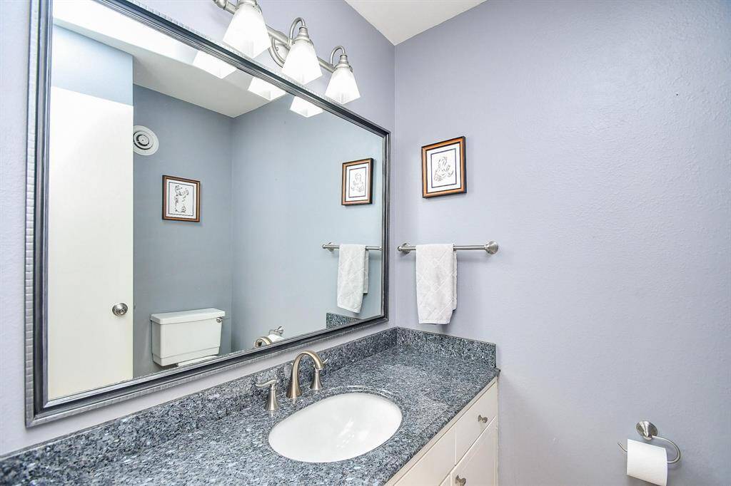 This bathroom features a modern design with light gray walls, a large mirror, and sleek lighting fixtures. It includes a granite countertop, undermount sink, and white cabinetry, offering a clean and inviting space.
