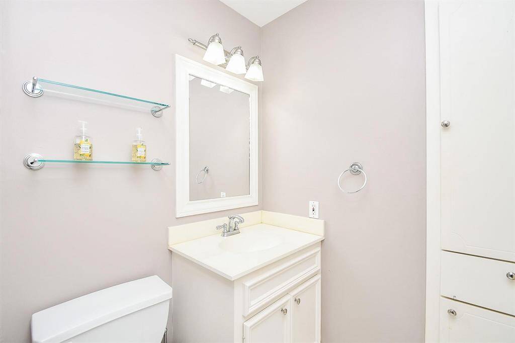 This is a bright, modern bathroom featuring a white vanity with a single sink, a sleek mirror, and overhead lighting. There are glass shelves for storage and a white cabinet for additional space, complemented by a neutral wall color.