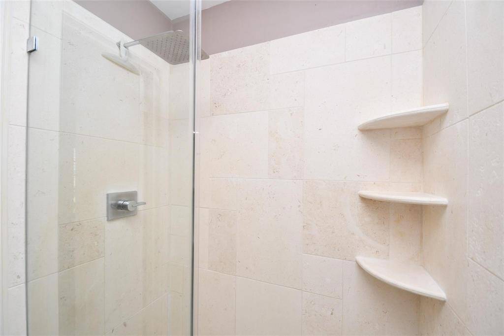 This photo shows a modern shower with beige tiled walls, a glass door, and built-in corner shelves. It features a square rain showerhead and a sleek metal handle for a clean, contemporary look.
