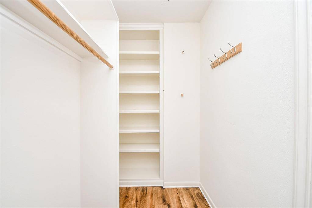 This photo shows a walk-in closet with white walls, built-in shelving, a wooden clothing rod, and a row of hooks on the wall, complemented by a wood-patterned floor. Perfect for organized storage.