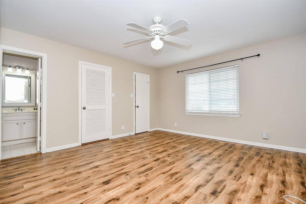 Seconday spacious room features vinyl wood flooring, neutral walls, and a ceiling fan. It has a large window with blinds and an adjoining bathroom with visible fixtures. Ideal for a bedroom or living space.