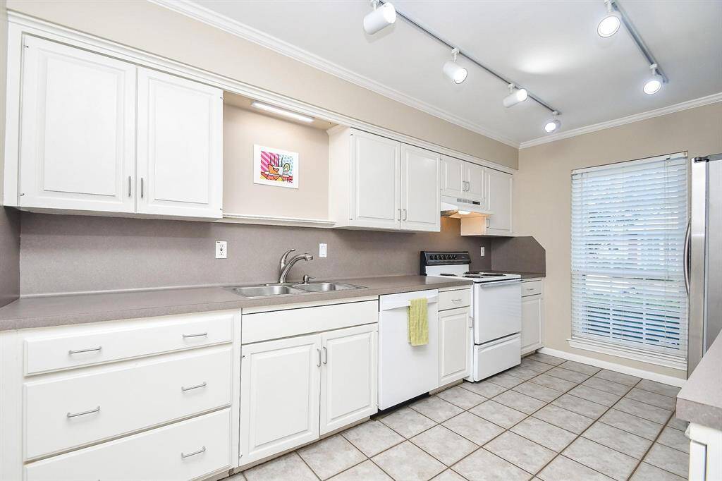 This kitchen features modern white cabinetry, ample counter space, and tiled flooring. It includes a stove, dishwasher, and a large window with blinds for natural light. Track lighting adds a contemporary touch.