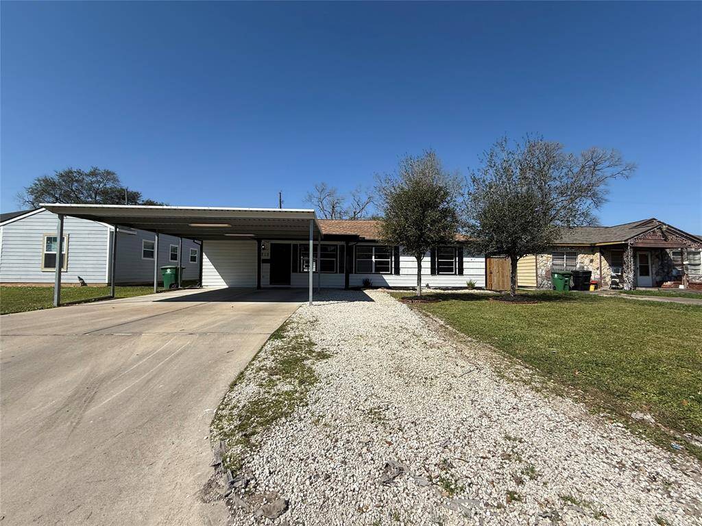 This photo shows a single-story home with a light-colored exterior and a two-car carport. The property features a gravel driveway, a small front lawn, and mature trees, offering a welcoming curb appeal under a clear blue sky.