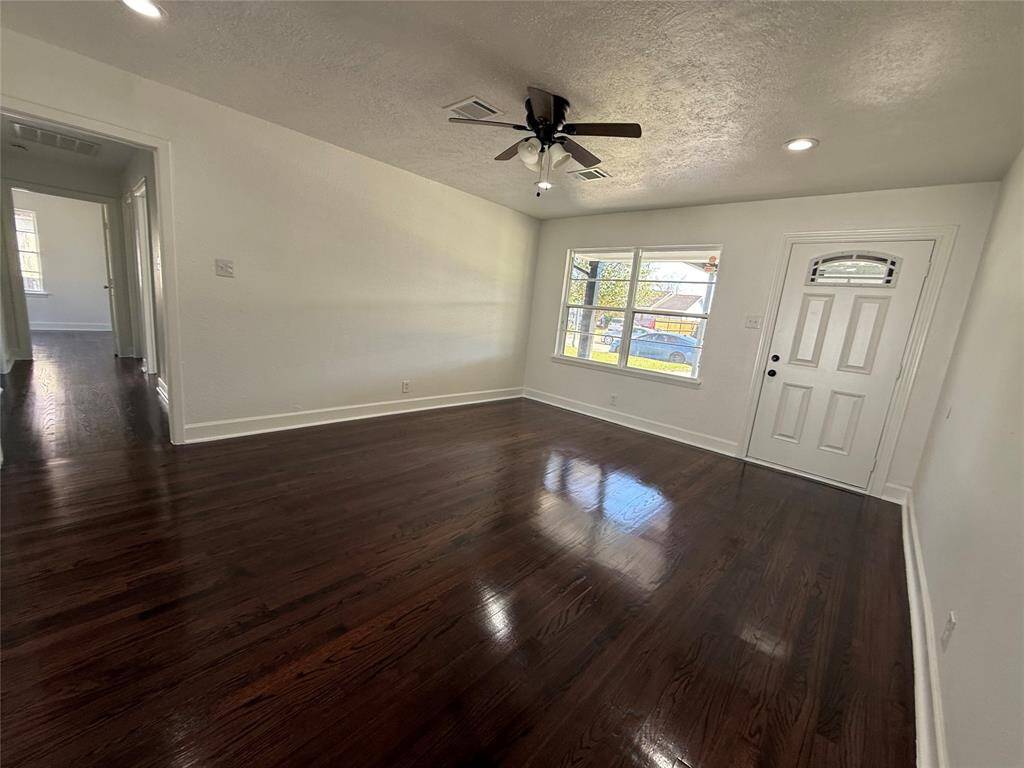 This photo showcases a bright living area with polished hardwood floors, neutral walls, and recessed lighting. It features a ceiling fan, a large window for natural light, and a welcoming front door. A hallway leads to additional spaces, creating an open and inviting atmosphere.