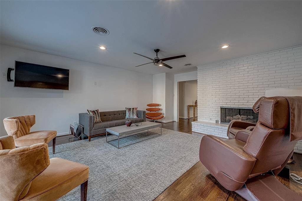 The large family room has a brick fireplace, wood floors and a ceiling fan.  You can see the doorway leads to the entry.